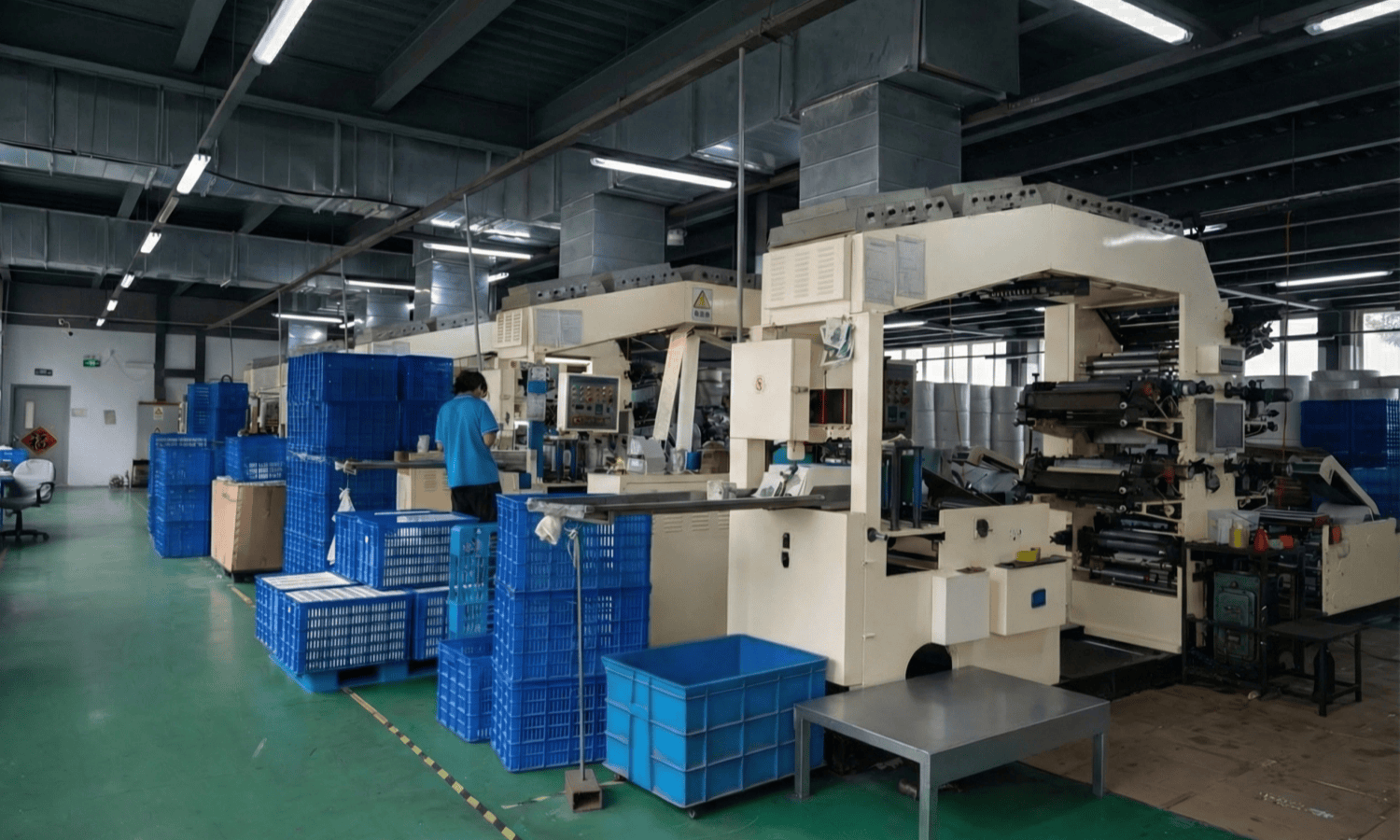 Clean and organized flexible packaging manufacturing plant interior, showing industrial rotogravure printing machines, stacked blue plastic crates, and a technician operating production equipment
