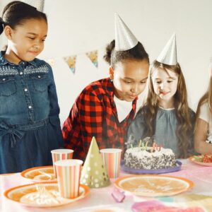 Group of diverse kids celebrating a birthday party with cake and party hats indoors.